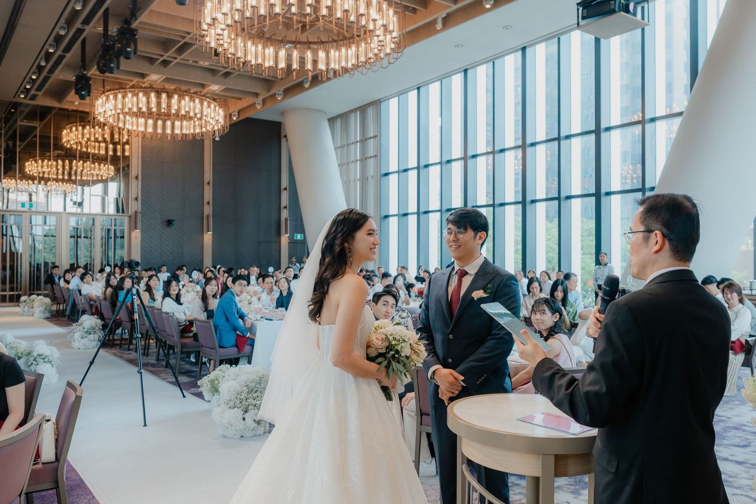 Wedding ceremony in a modern venue with floor-to-ceiling windows and elegant chandeliers.