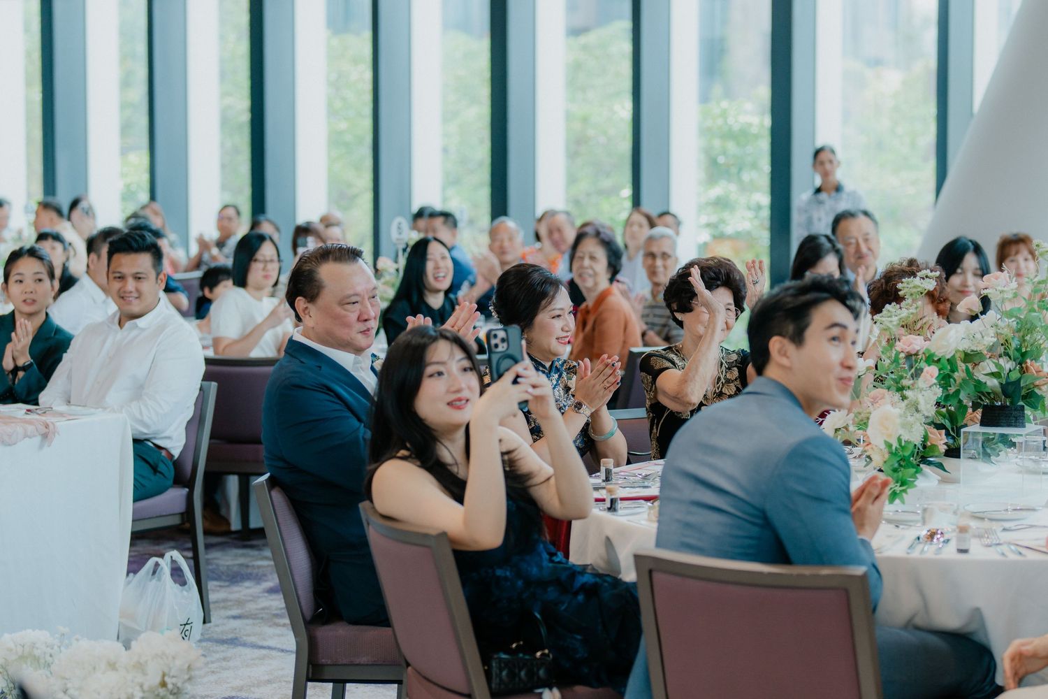 Wedding reception guests seated at decorated tables in a bright modern venue with large windows.