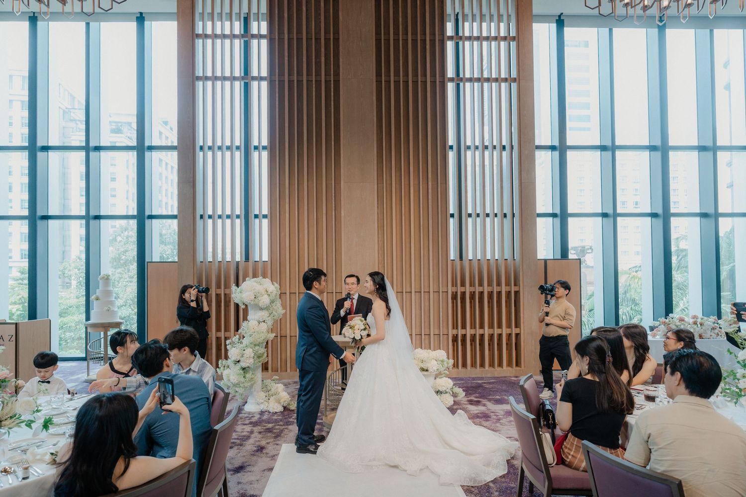 Wedding ceremony in a modern venue with floor-to-ceiling windows as guests look on.