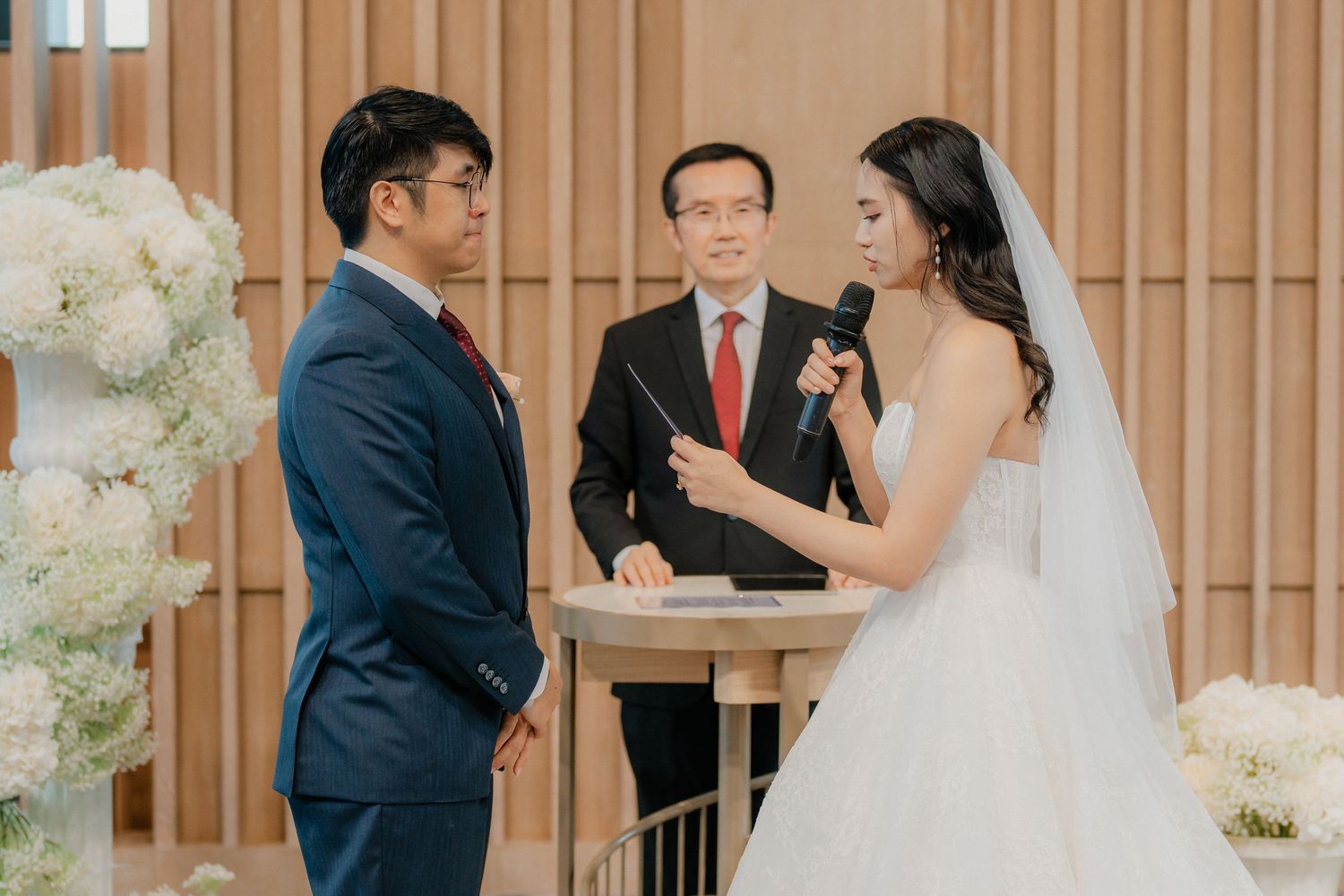 A wedding ceremony in progress with a bride in white gown and groom in navy suit at a modern venue with floral decor.