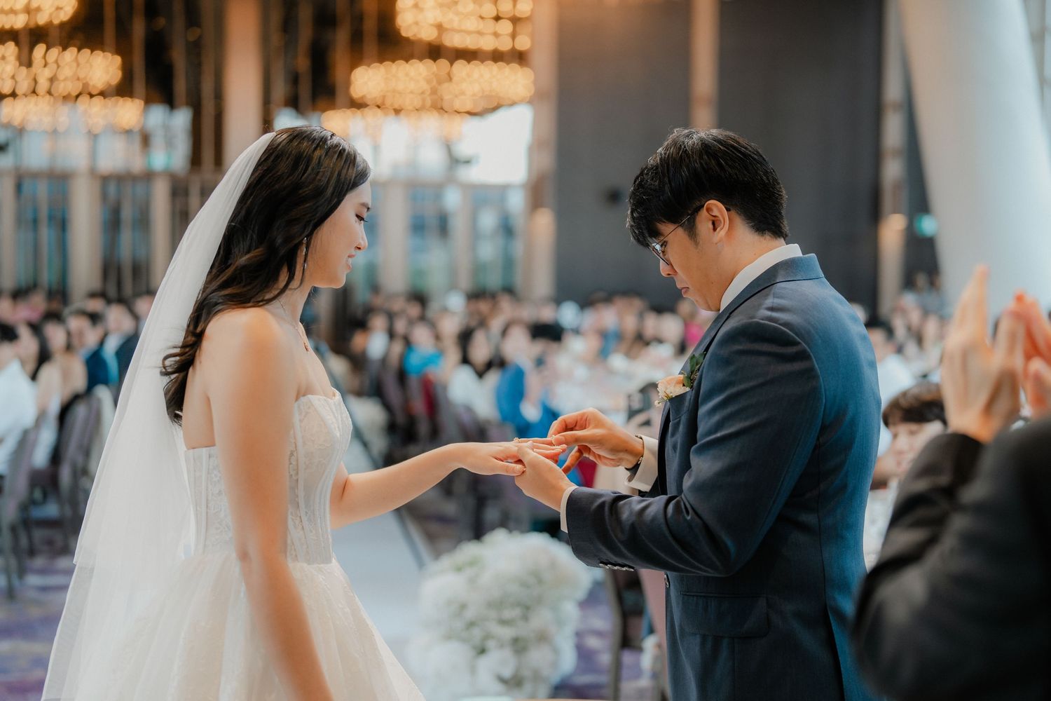 Exchange of rings during wedding ceremony with guests looking on in background.