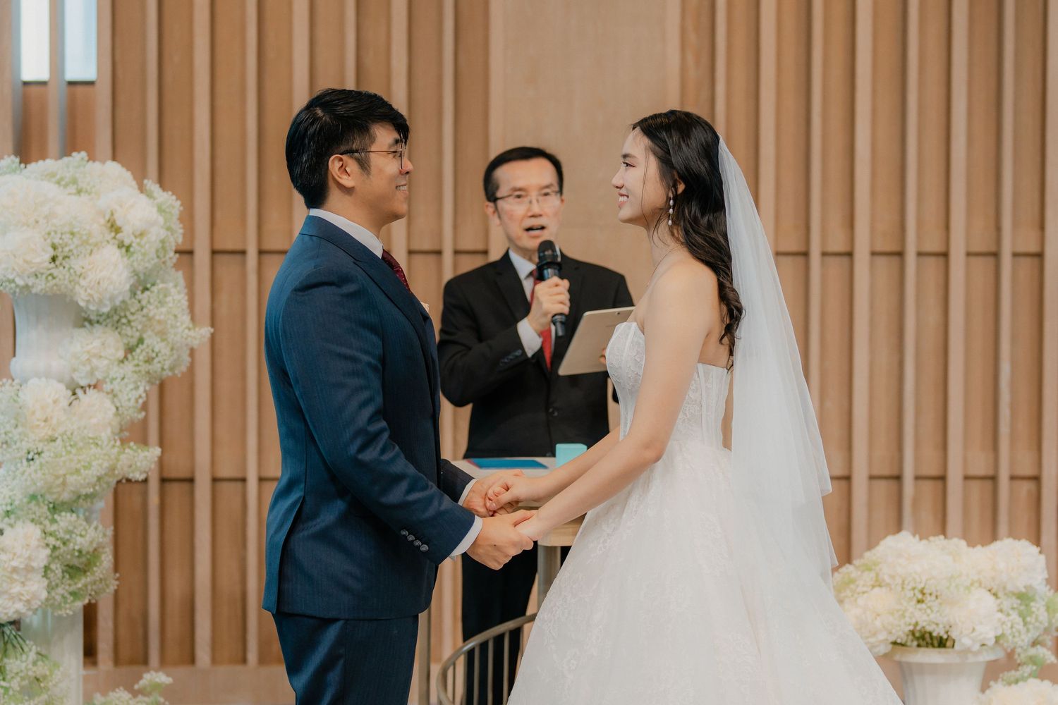 Wedding ceremony with officiant and couple standing in front of ornate wood-paneled wall with white flowers.