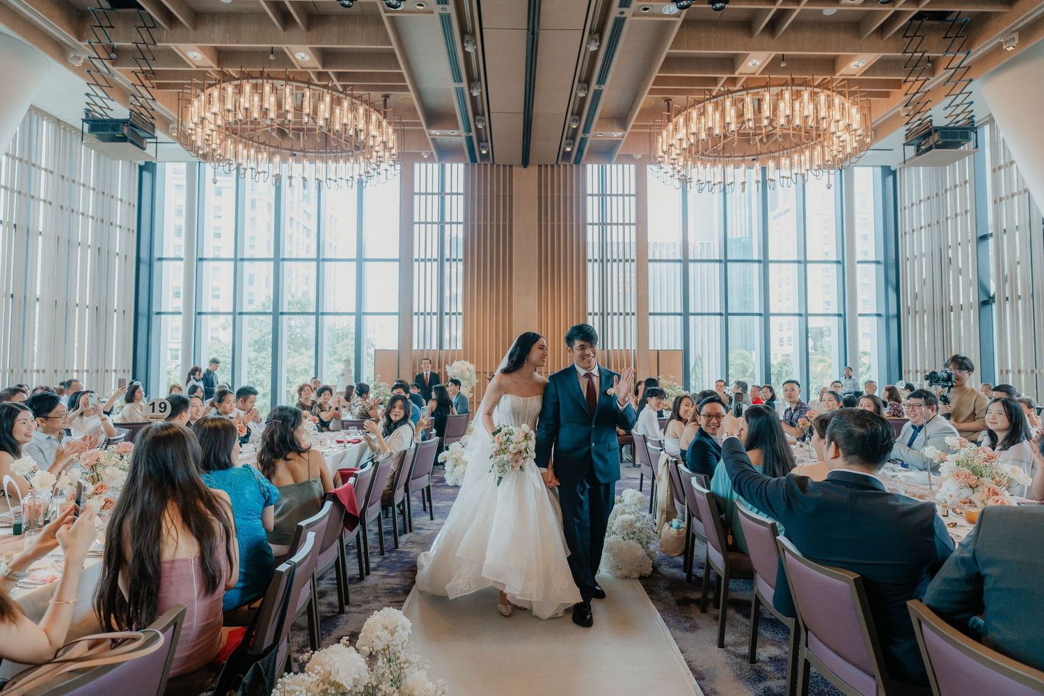Newlyweds walking down the aisle while guests applaud in an elegant venue with city views.