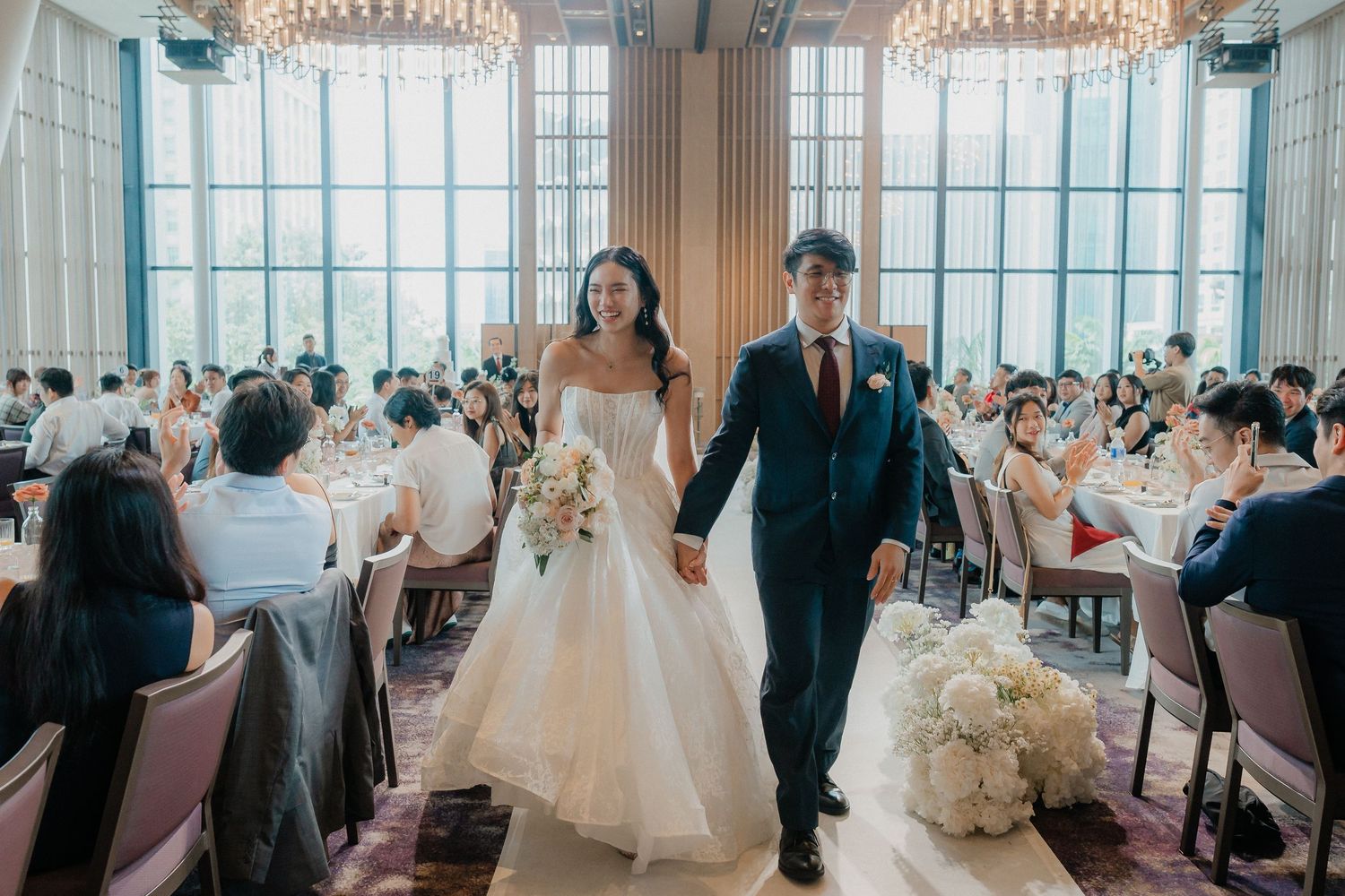 A couple walks down an aisle in an elegant ballroom with large windows and crystal chandeliers.