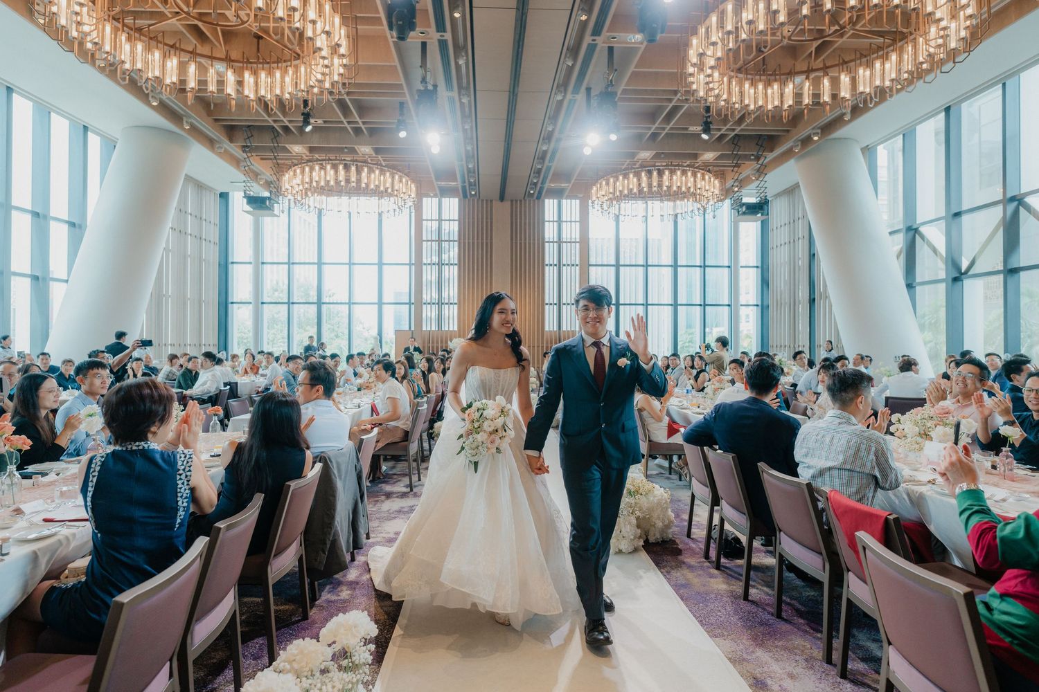 Couple walks down aisle at elegant wedding reception in modern venue with floor-to-ceiling windows and crystal chandeliers.