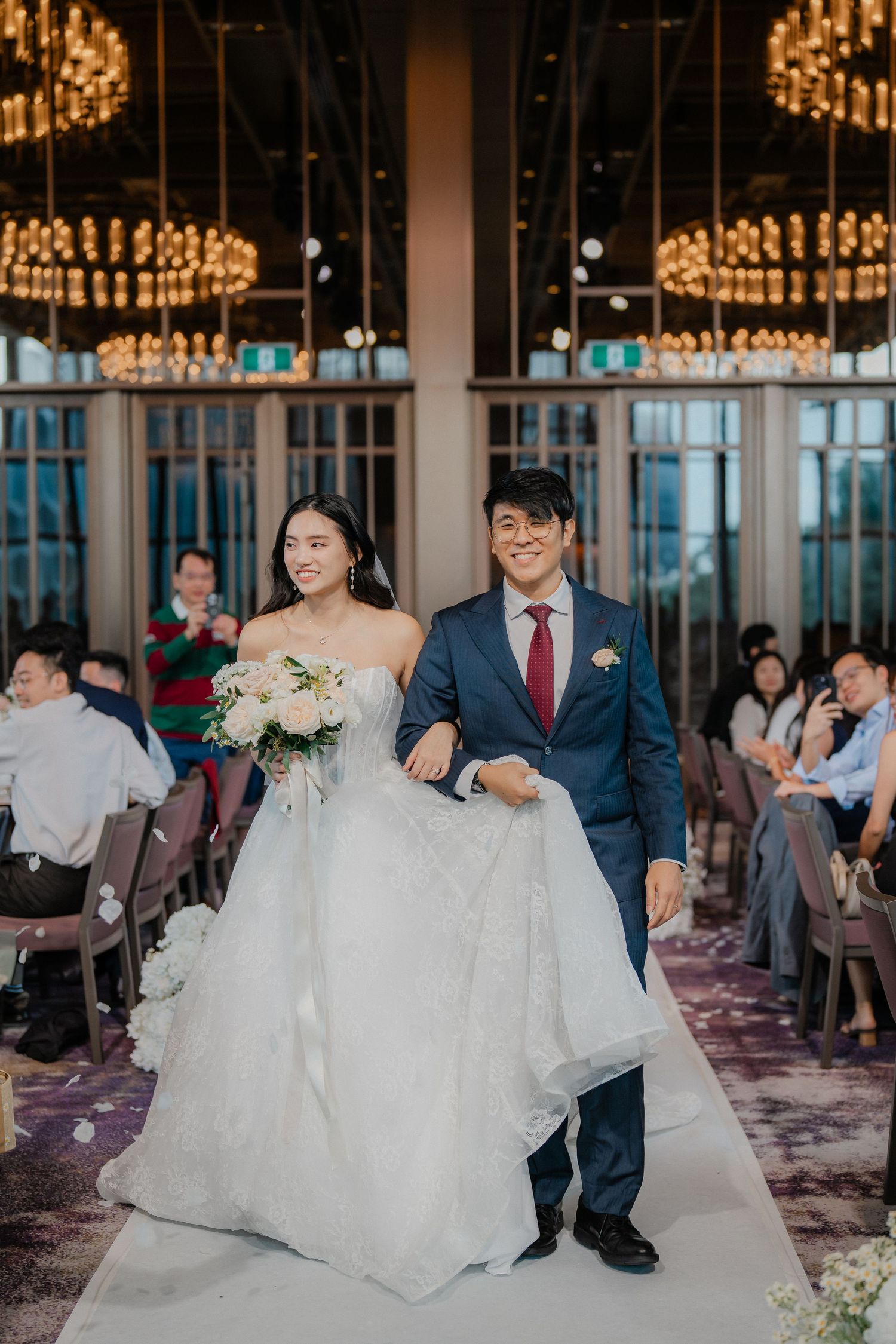 Newlyweds walk down aisle in elegant reception hall with crystal chandeliers.