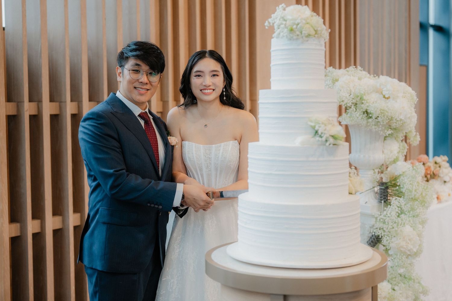 Wedding couple and guests pose with an elegant white tiered wedding cake adorned with floral decorations.