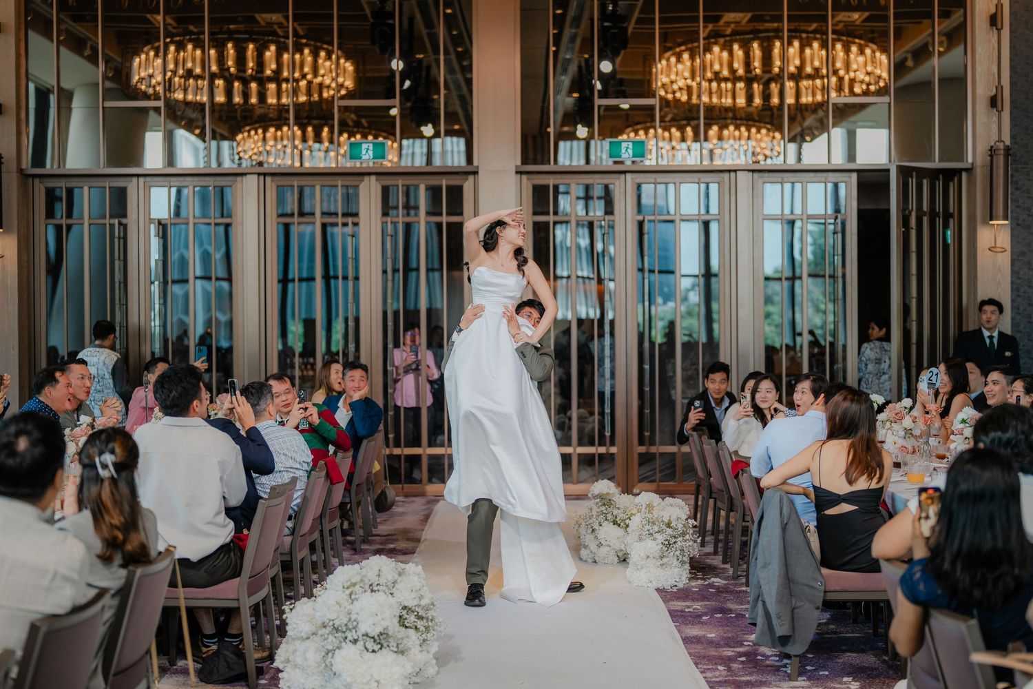 Wedding celebration in a modern venue with tall chandeliers and full-length windows as guests watch from their seats.