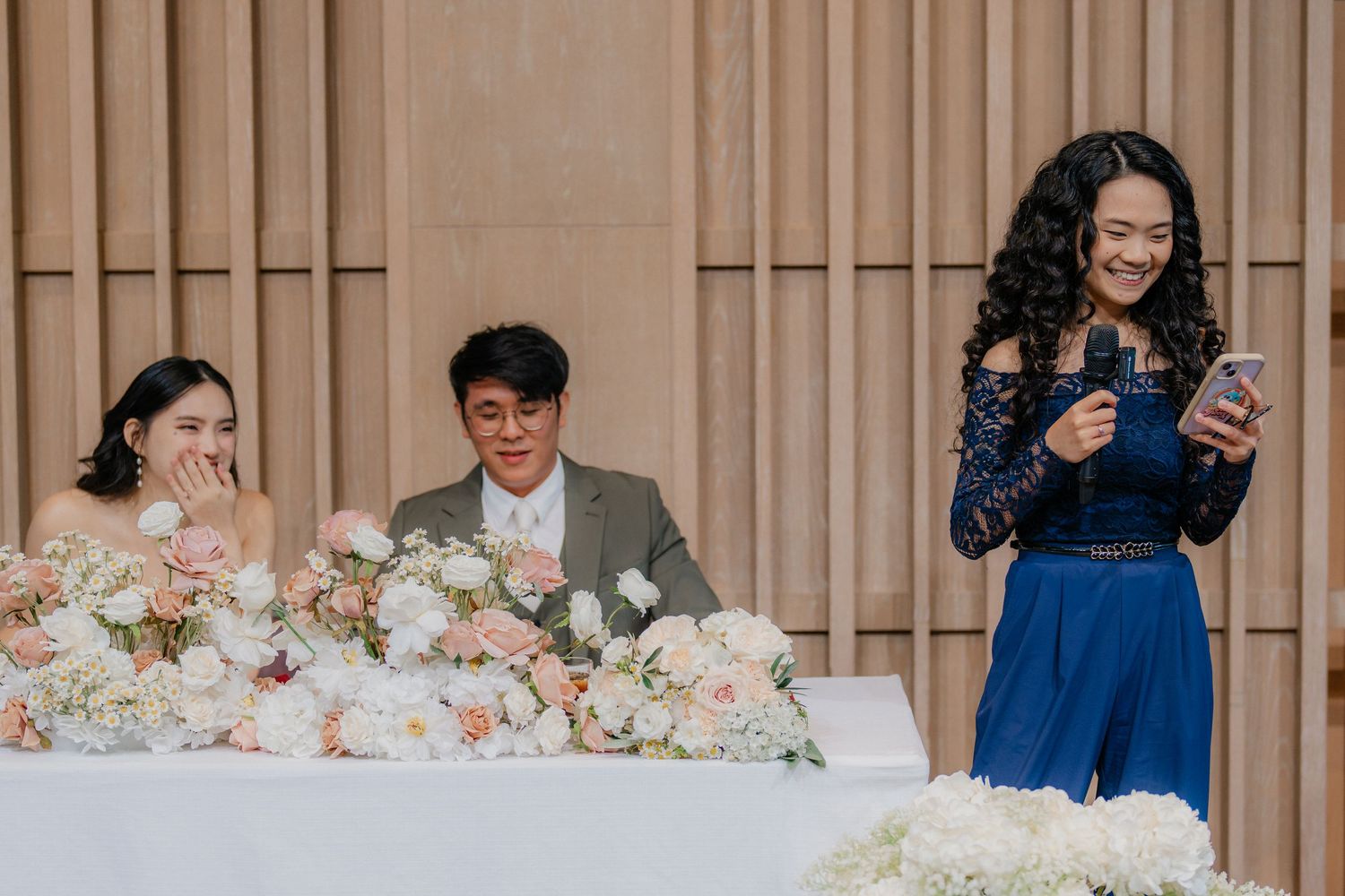 Wedding reception at head table with floral arrangements and guest in blue dress using phone.