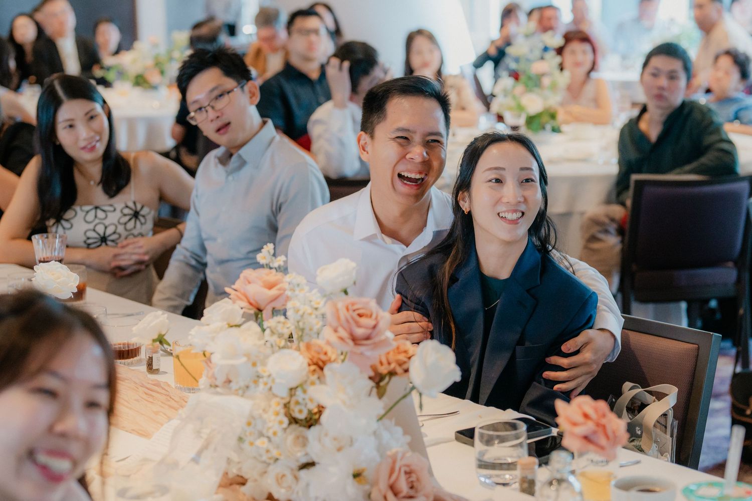 Wedding guests laugh joyfully at a reception dinner table decorated with floral centerpieces.