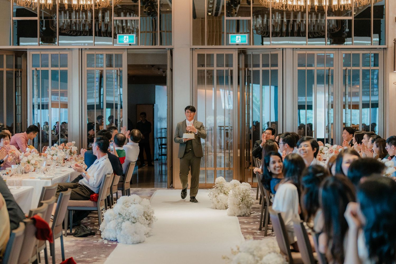 People seated at tables in a bright ballroom watching a speaker during a wedding reception.