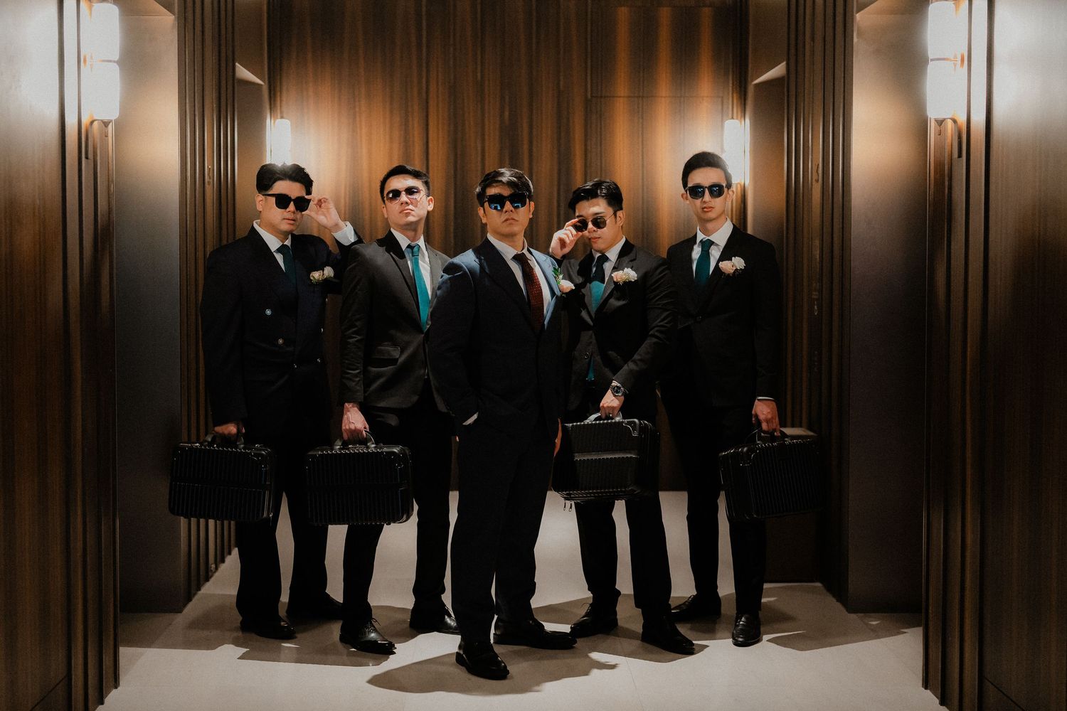 A group in matching dark suits and sunglasses pose together in an elevator lobby area.