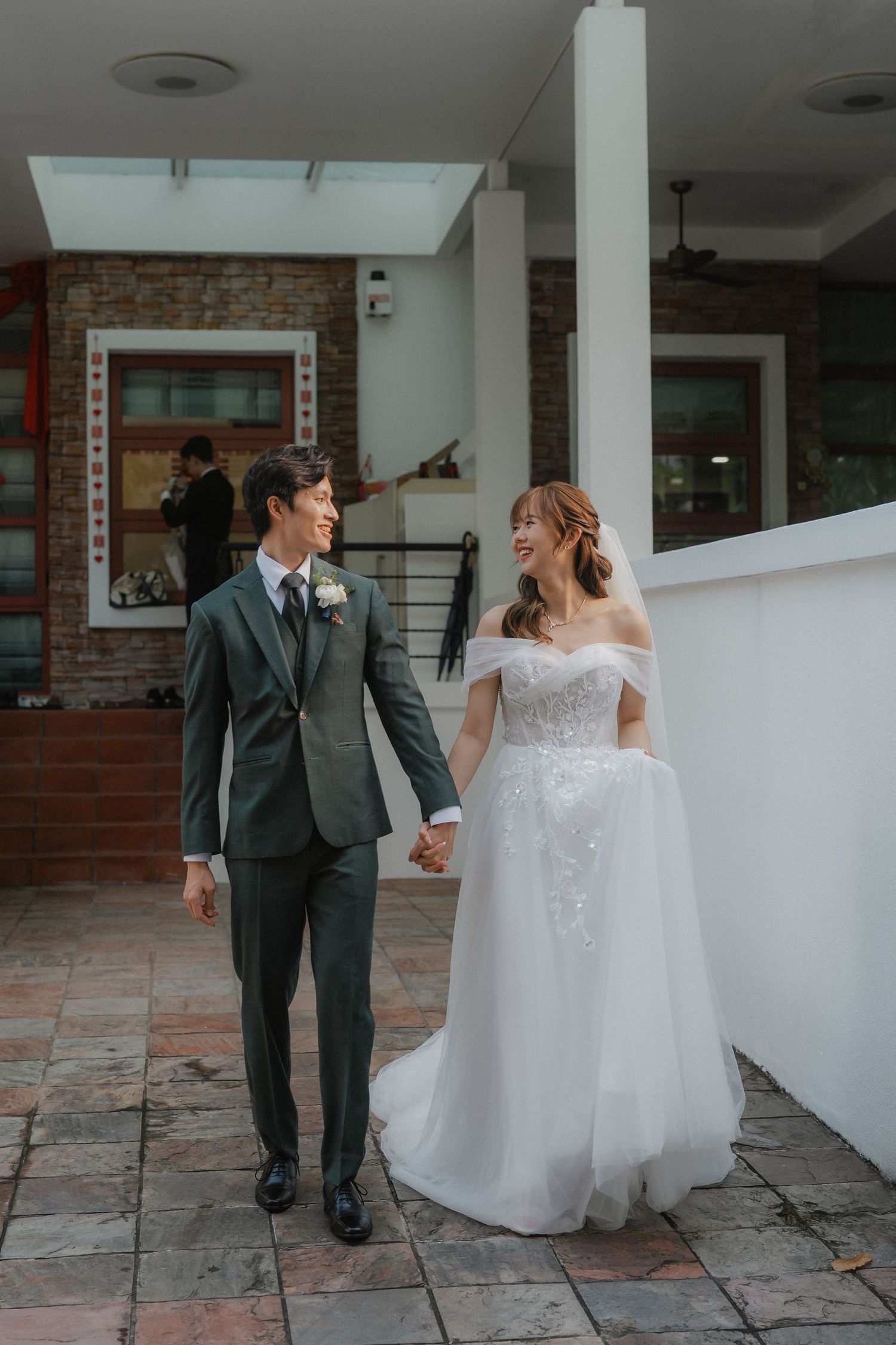 A wedding couple walks hand in hand on a brick patio outside a building with rustic brick walls and white columns.