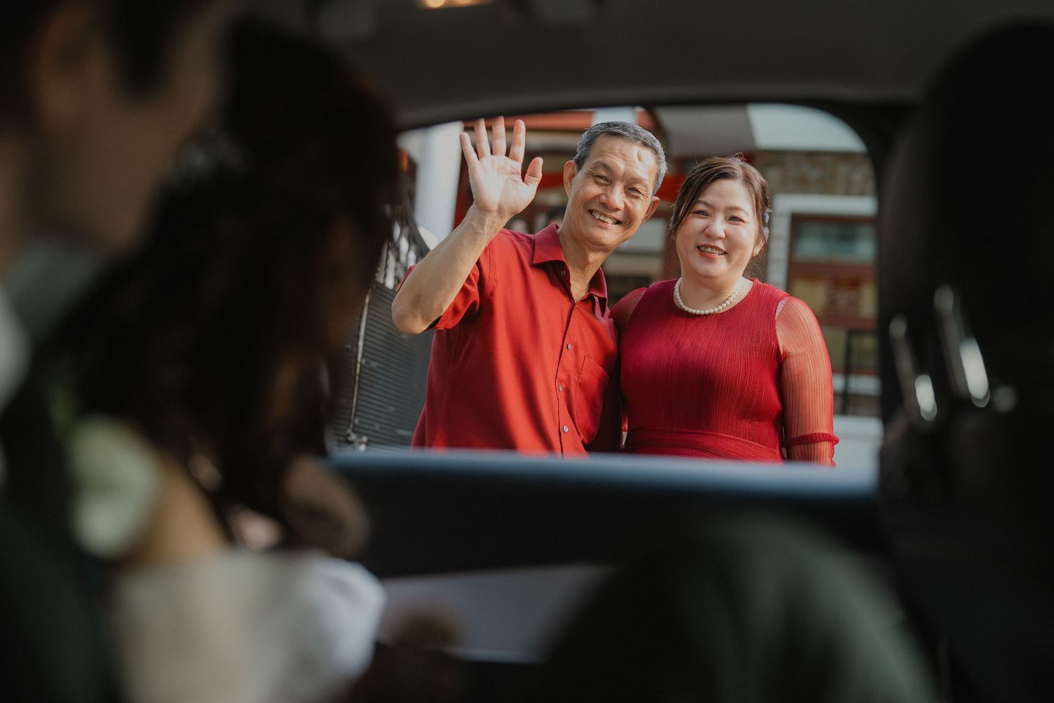 A couple in red outfits wave through a car window during a wedding celebration.