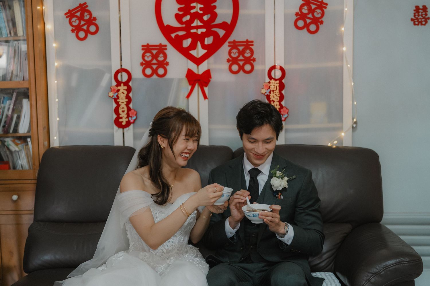 Newlyweds sit on couch looking at phones with traditional Chinese wedding decorations in background.