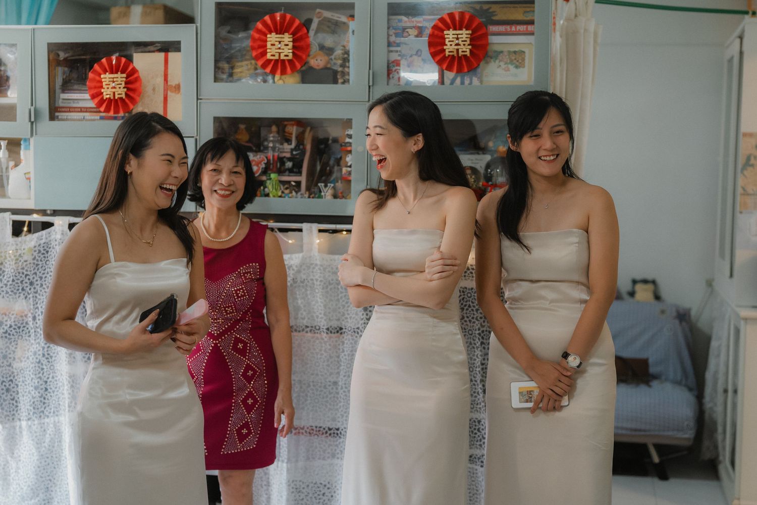 Bridesmaids in cream-colored dresses share a joyful moment with someone in red during wedding preparations.