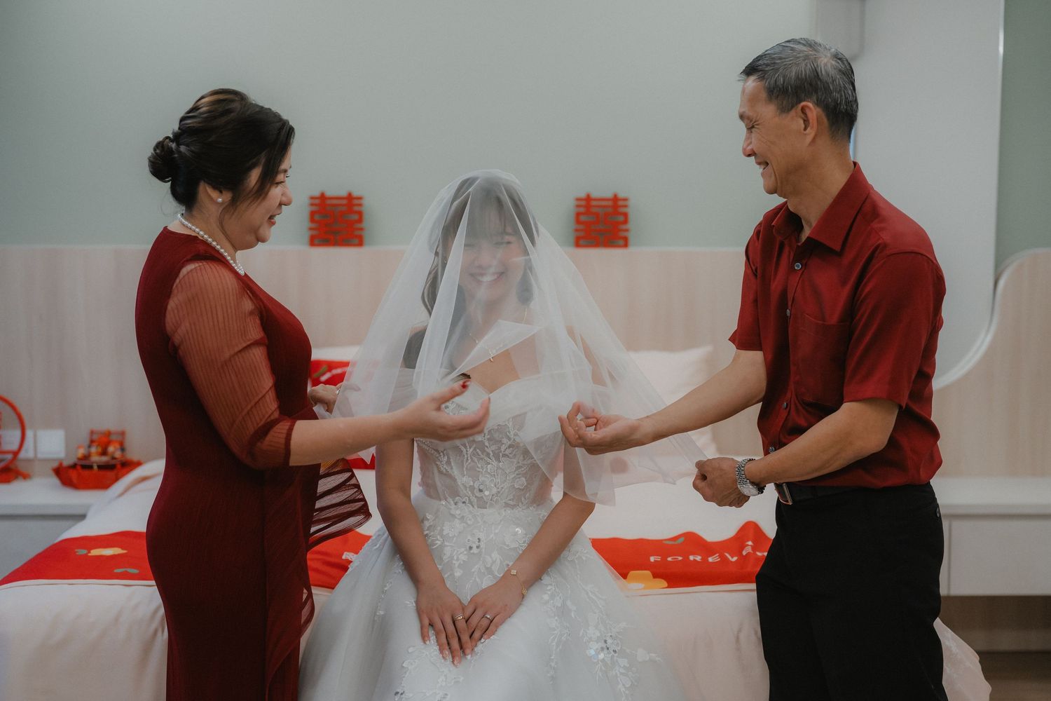 Traditional wedding preparation scene with parents helping adjust bride's veil against white walls with red decorations.