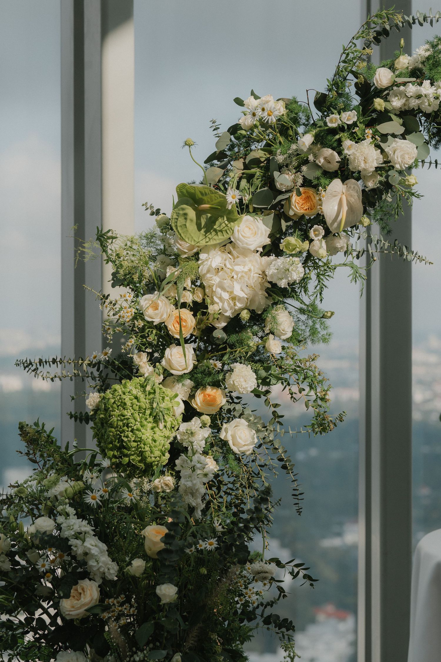 Elegant white floral arrangements with roses and greenery decorate a modern glass-walled wedding venue with city views.