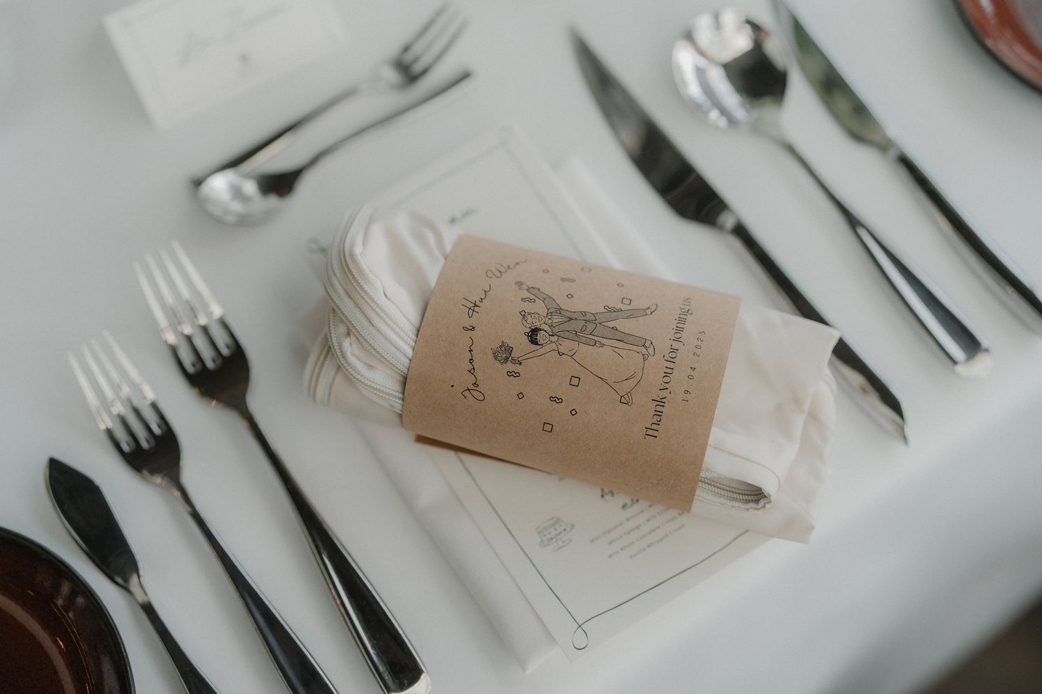 Wedding place setting with silverware and rustic brown favor tag on white tablecloth.