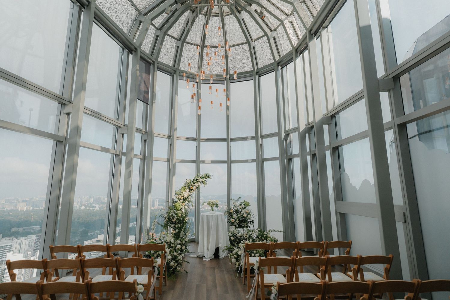 A modern glass-domed wedding ceremony space with wooden chairs arranged in rows and floral decorations.