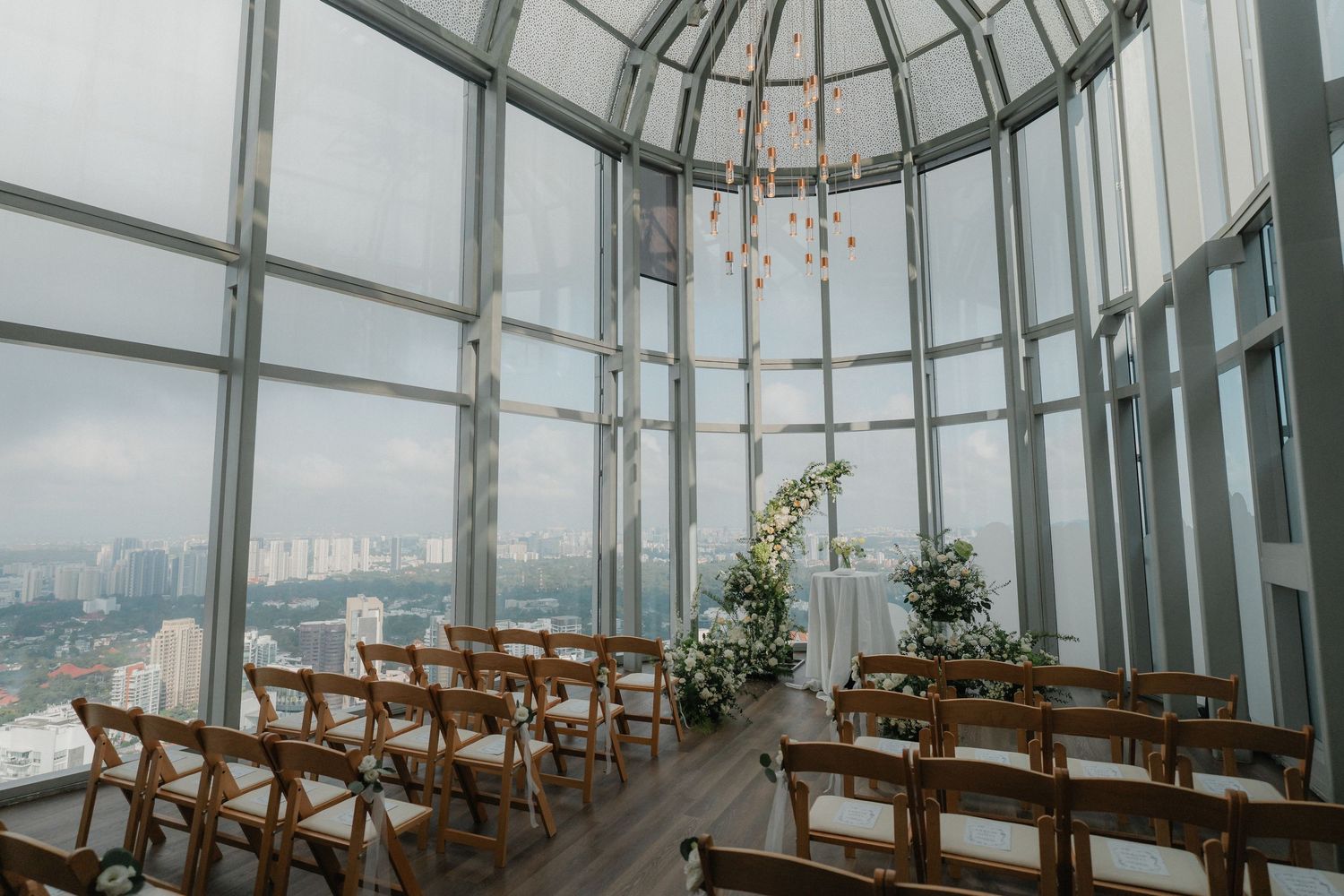Modern glass-domed wedding ceremony space with wooden chairs and floral decorations.