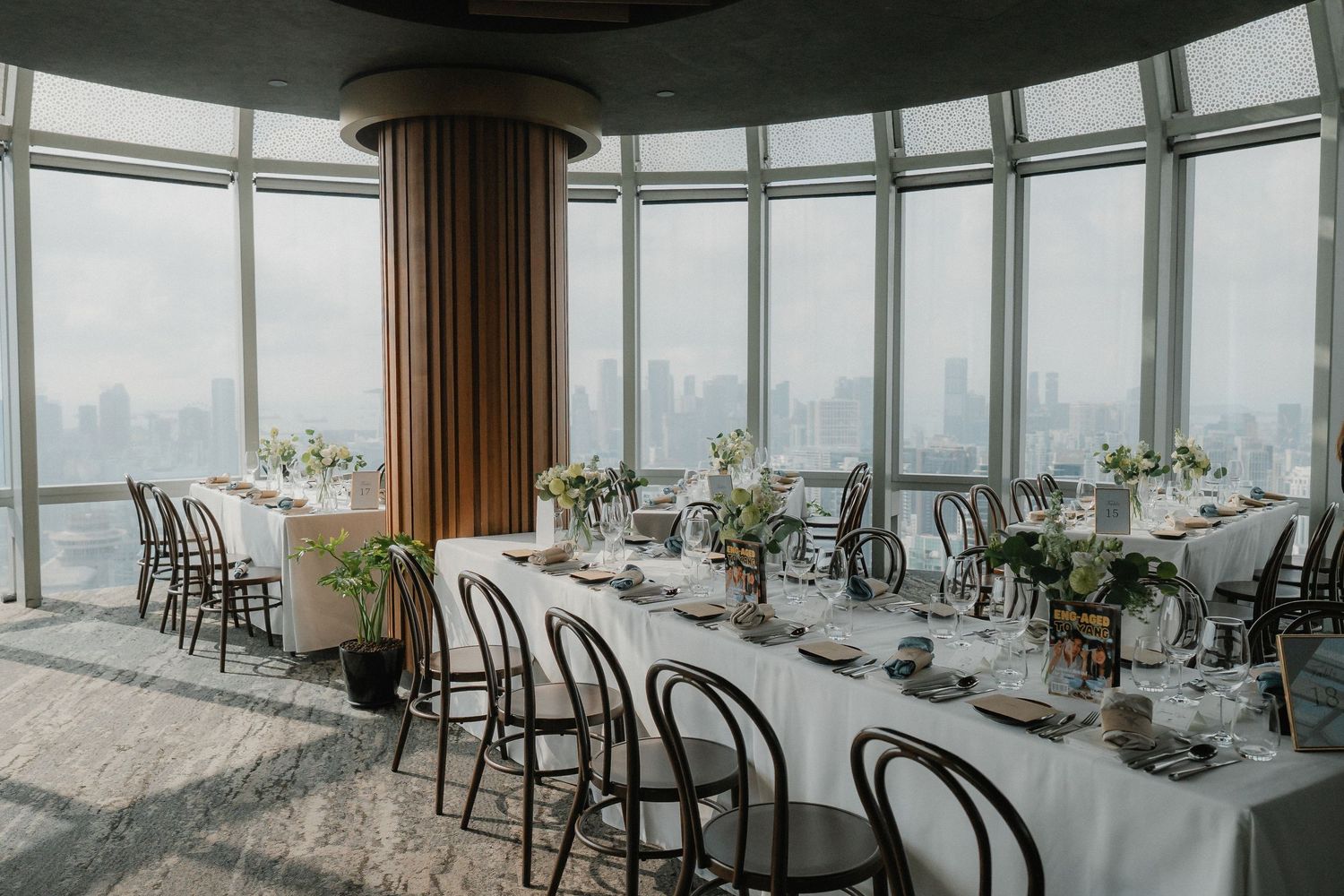 A sophisticated dining setup with bentwood chairs and long tables in a glass-walled venue overlooking a city.
