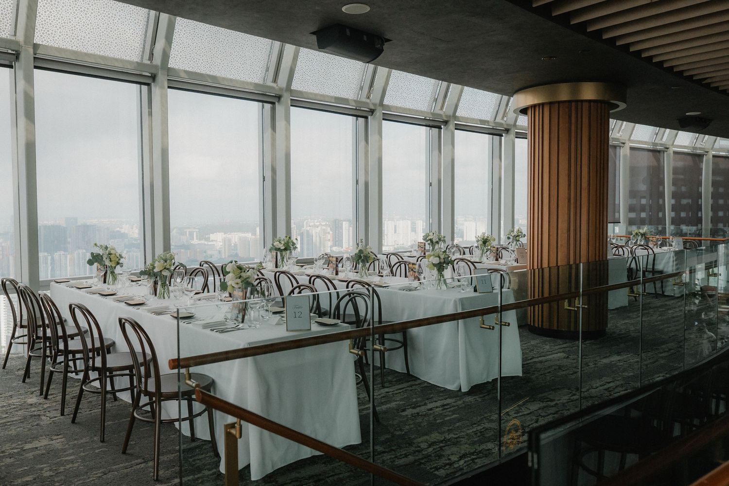 Elegant wedding reception venue with panoramic windows, long white-clothed dining tables and bentwood chairs.