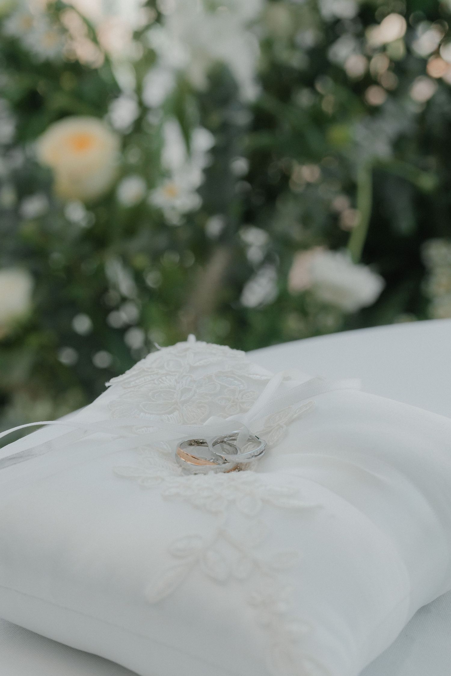 Wedding rings rest delicately on a white embroidered cushion with blurred floral background.