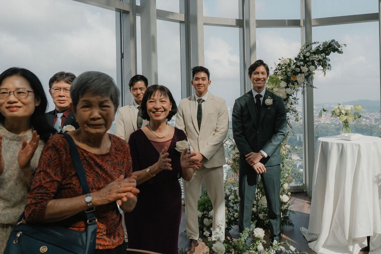 Wedding guests gather in bright venue with floor-to-ceiling windows and city views.