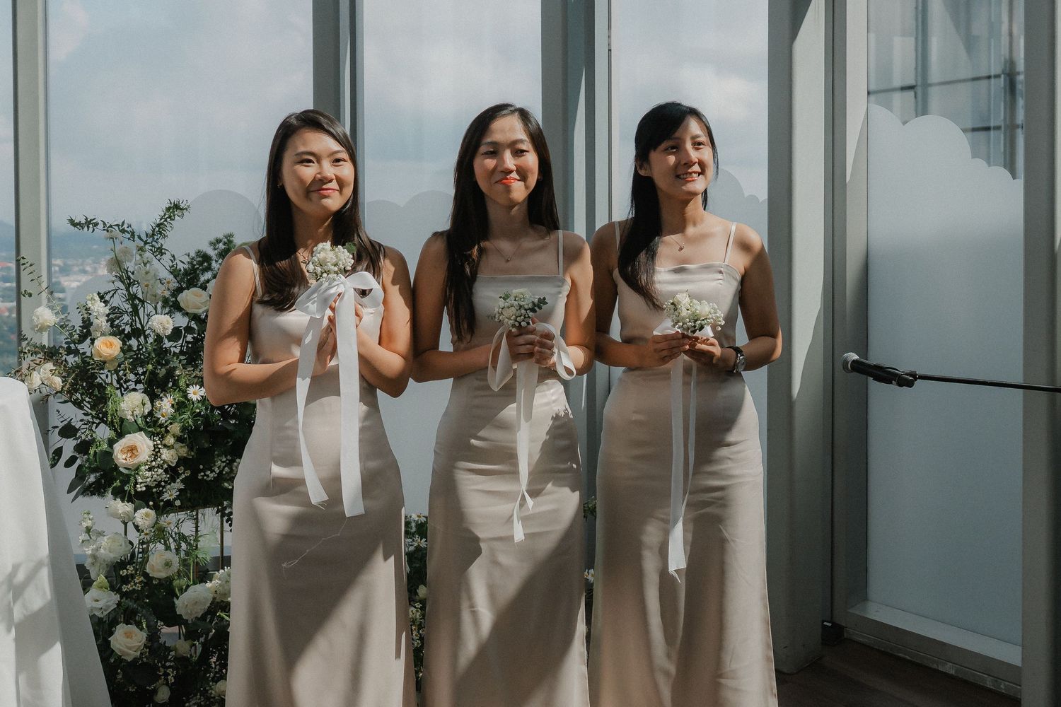 Bridesmaids in elegant white dresses pose by large windows in modern venue.