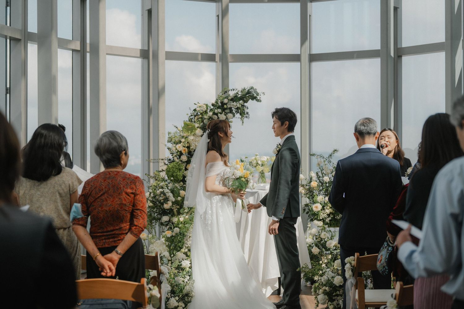 Wedding ceremony takes place in a modern glass-walled venue with floral arch decorations and guests in attendance.