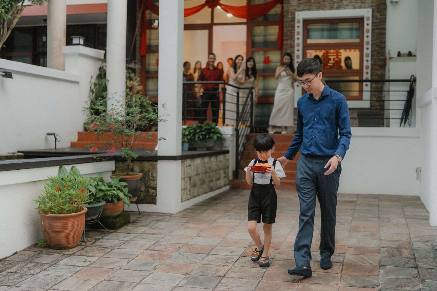 A groom in a blue shirt walks with a young ring bearer on a terrace while guests look on in the background.