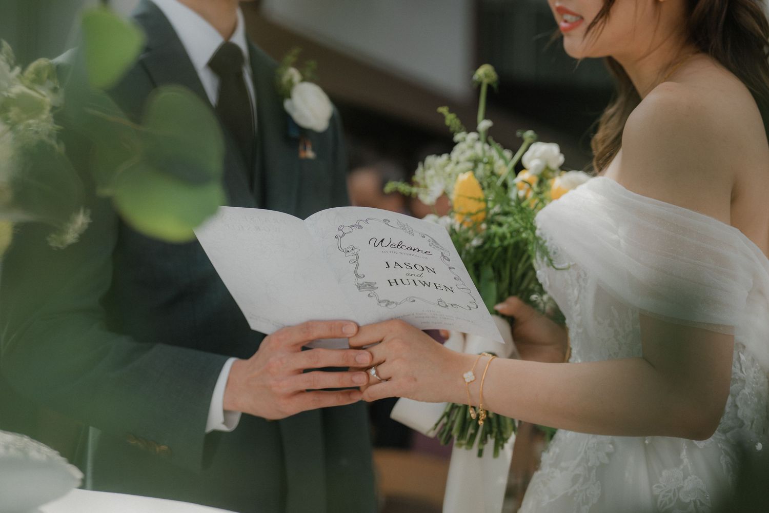 A couple reads wedding vows while holding a bouquet of yellow and white flowers during an intimate ceremony.