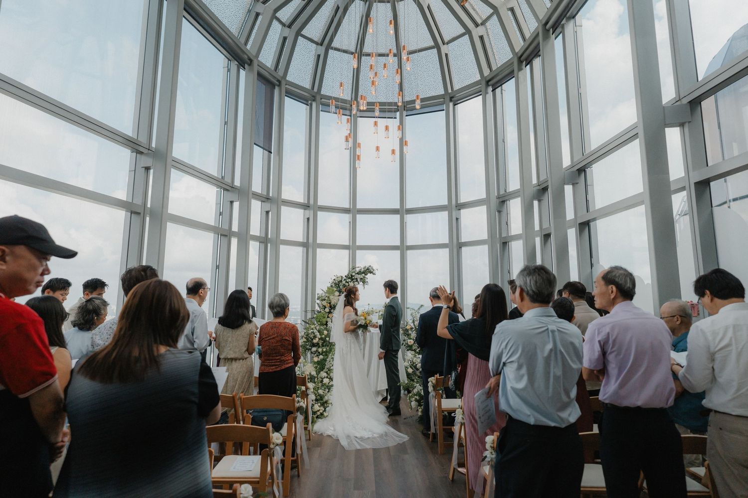 Wedding ceremony in modern glass atrium with guests seated beneath domed ceiling.