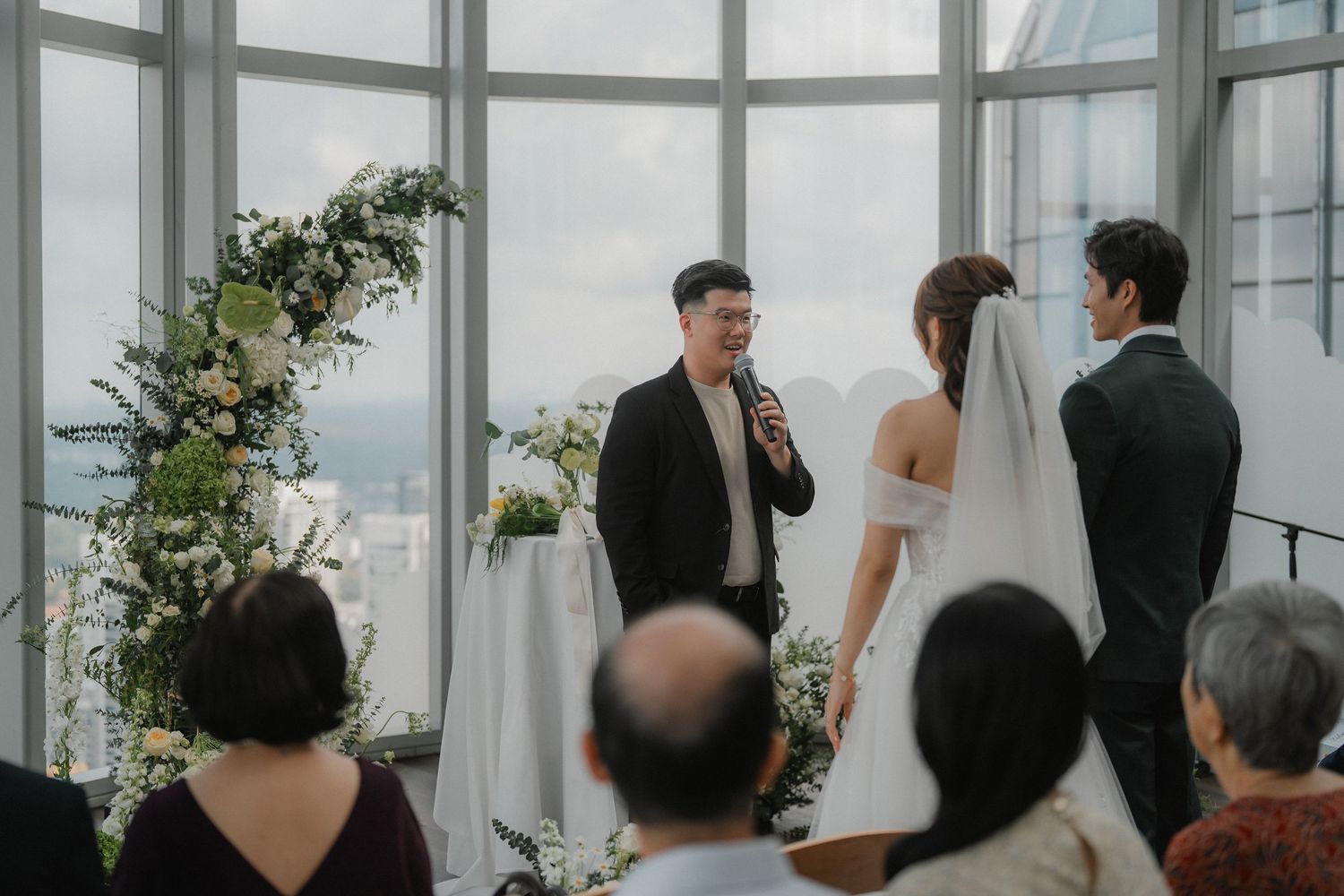 Wedding ceremony taking place in a modern venue with floor-to-ceiling windows and floral decorations.