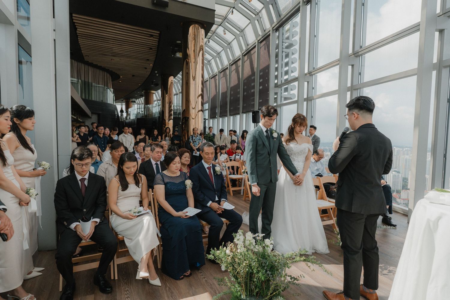 Guests seated in rows watch a wedding ceremony in a modern glass-walled venue with city views.