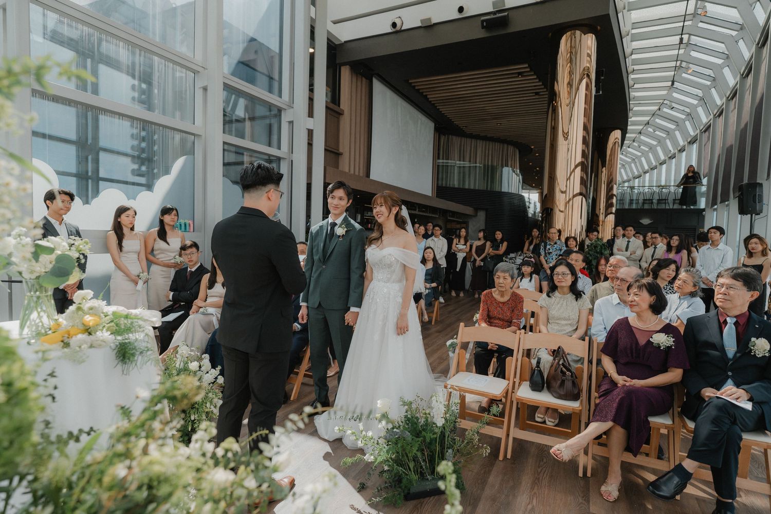 Wedding celebration in bright conservatory-style venue with natural light and floral decorations.