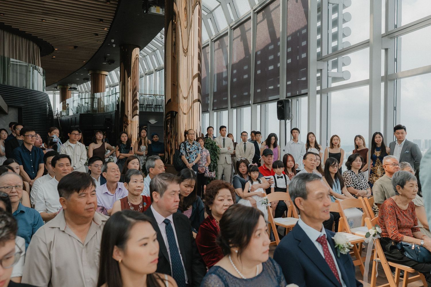 Wedding ceremony guests seated in modern venue with floor-to-ceiling windows.