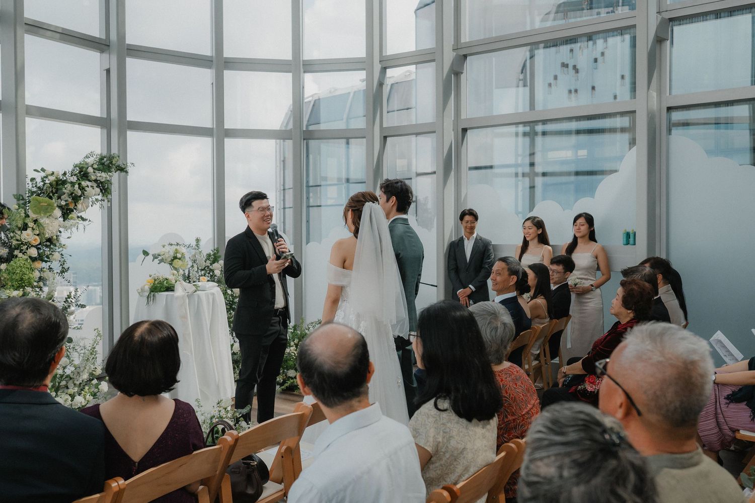 Wedding guests seated in rows at a modern venue with floor-to-ceiling windows and floral decorations.