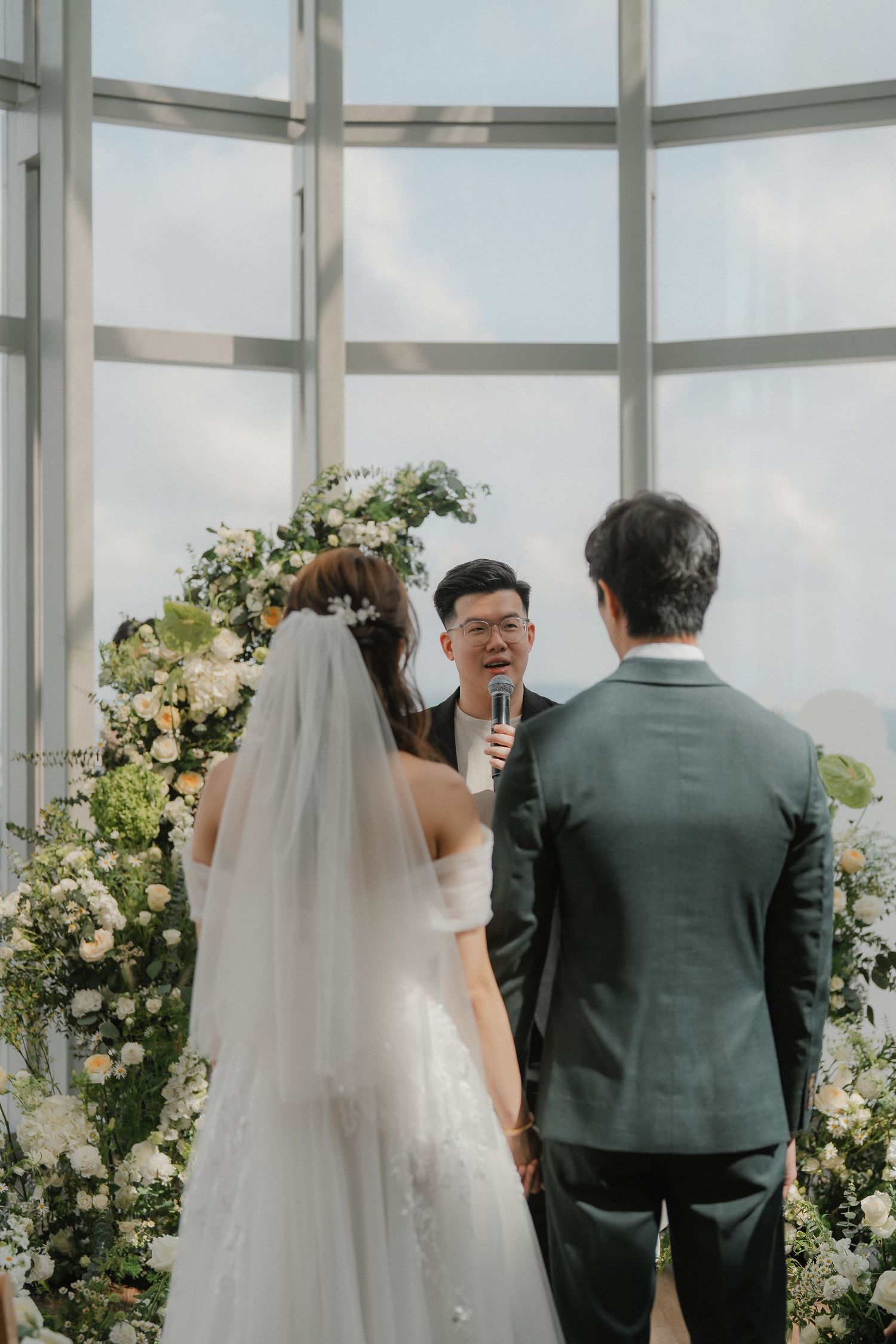 A wedding ceremony in a bright glass-walled venue with floral arch decorations.
