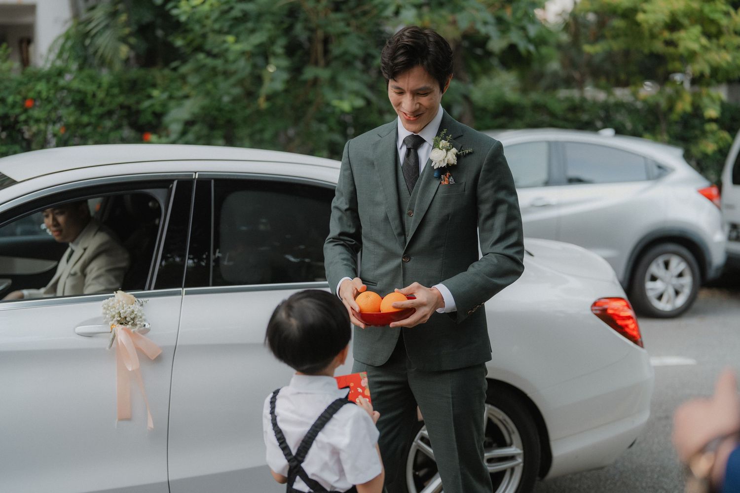 Groom in a dark suit holds something while standing next to a decorated car during a wedding ceremony.