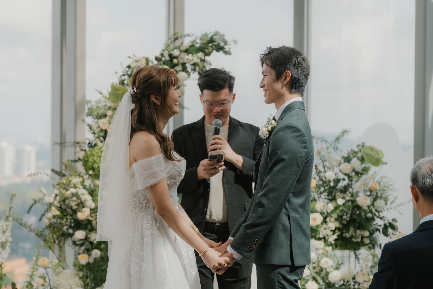 Wedding ceremony taking place in bright sunlit venue with floral arch and city views.