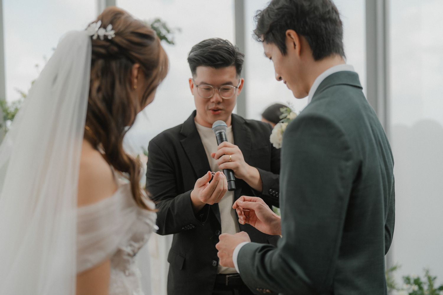 Wedding ceremony couple exchanging rings with officiant holding microphone during intimate outdoor ceremony.