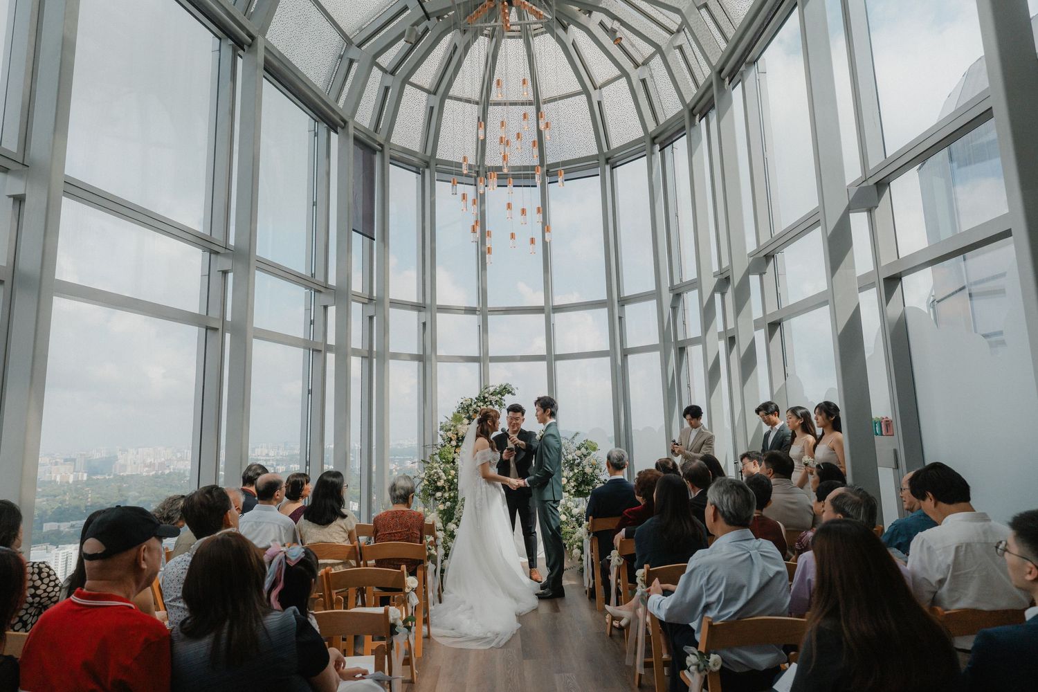 Wedding ceremony setup in a glass-domed space with modern chandeliers and elegant decor.