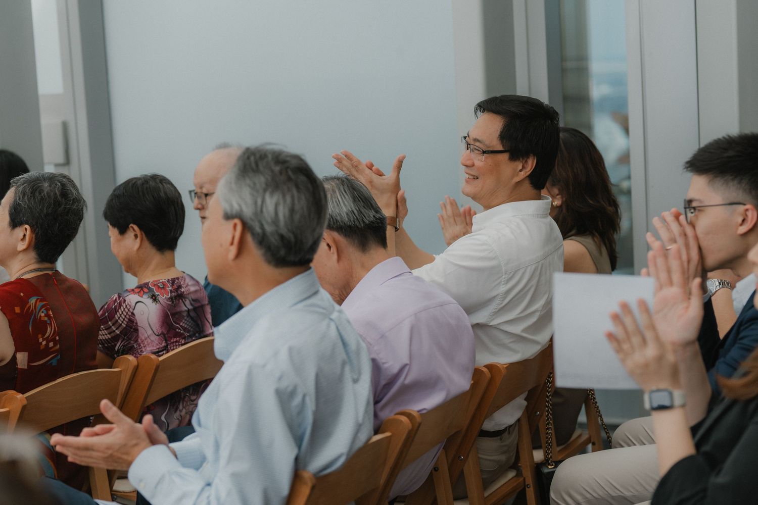Wedding guests seated in wooden chairs indoors watching a ceremony presentation.