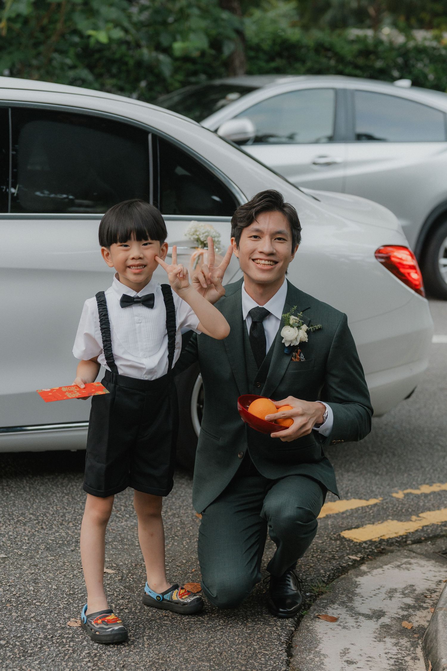 A boy in suspenders and bow tie poses with a wedding attendant in a dark suit next to a parked car.