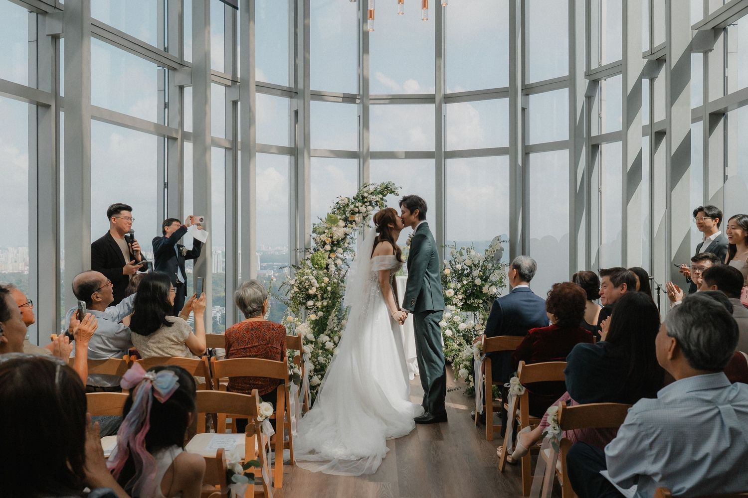 Panoramic view of a wedding ceremony in a glass-enclosed venue with wooden benches and floral decorations.