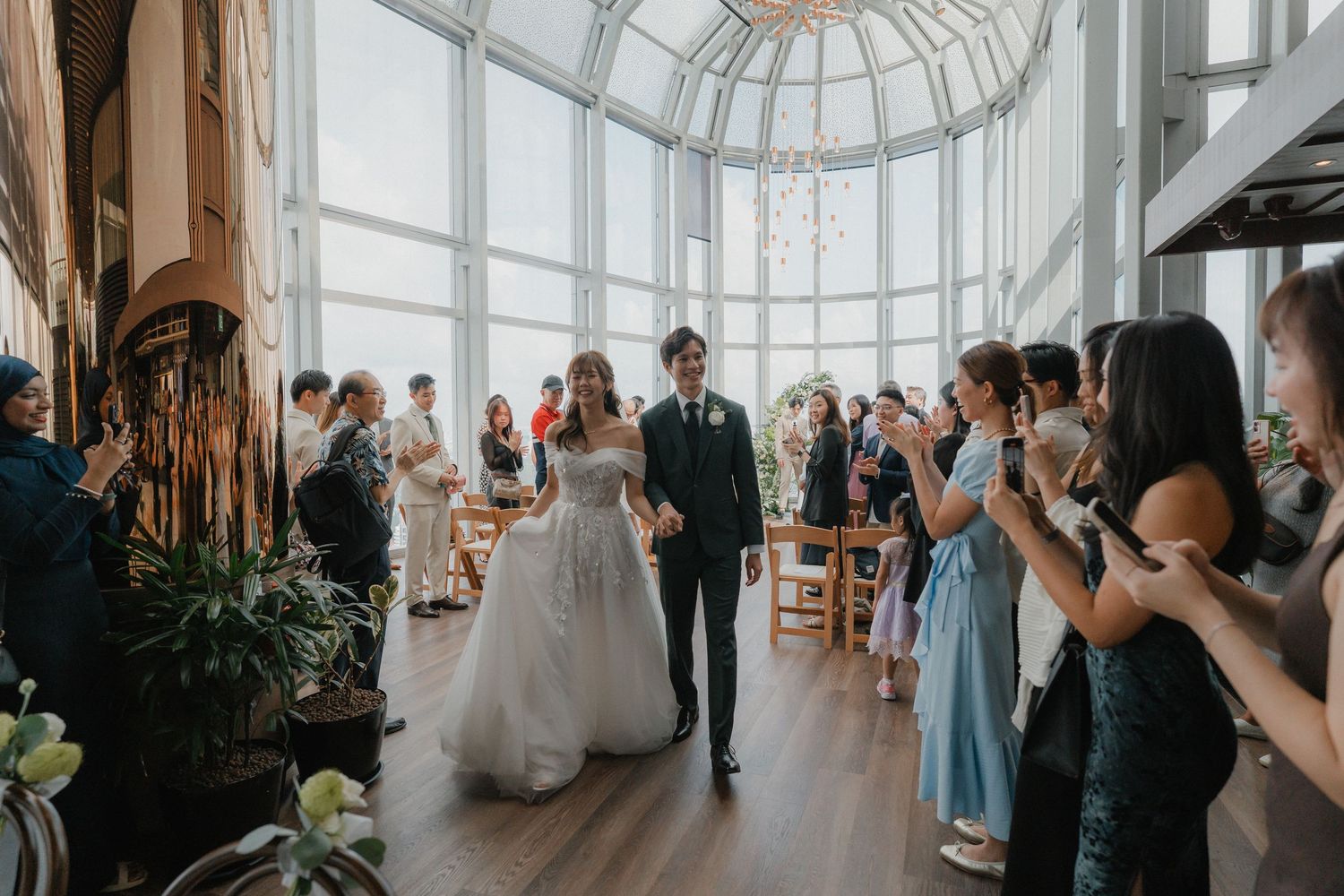 Wedding ceremony in progress inside a modern glass venue with panoramic city views.