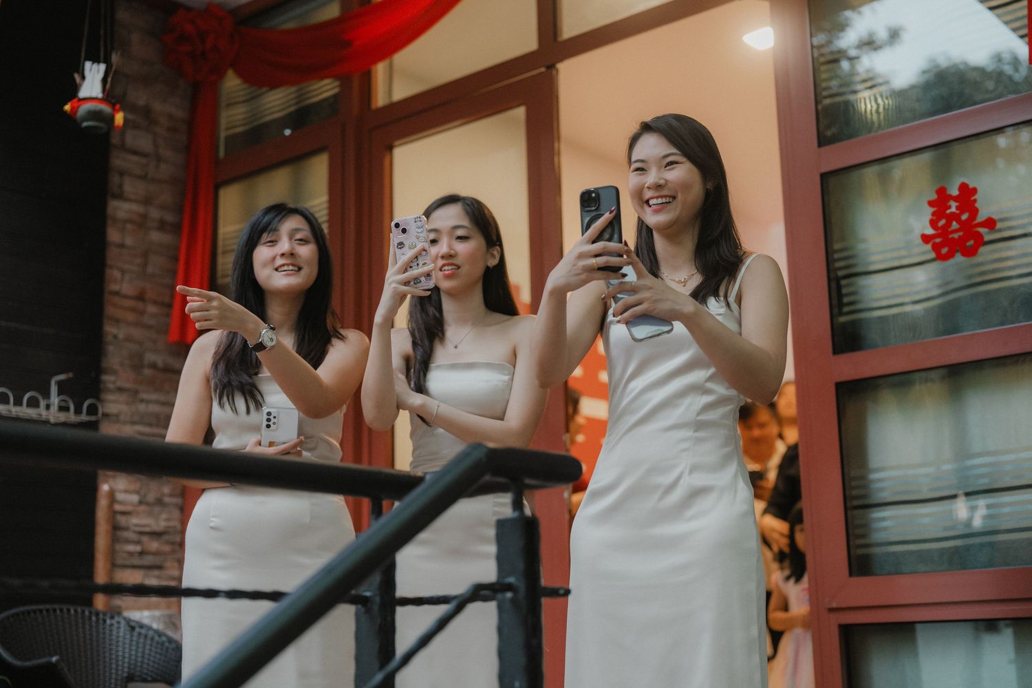 Three people in white dresses smile and take photos from a balcony with red decorations.