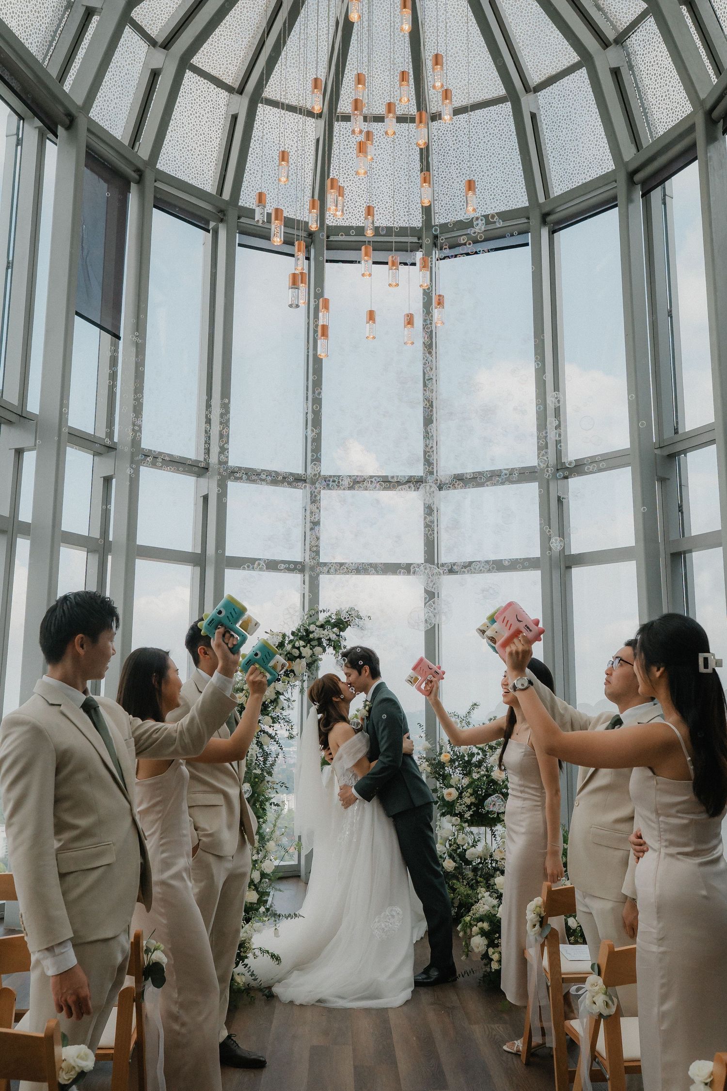 Wedding party dancing under a glass dome ceiling with dramatic lighting and city views.